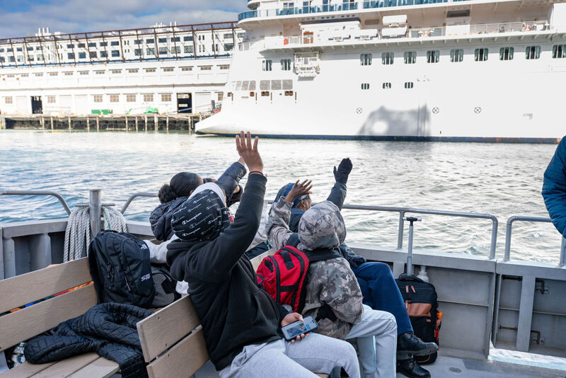The image shows a group of people on a boat, with some of them raising their hands. In the background, there is a large cruise ship docked at a pier. The sky is cloudy, and the water appears calm. The people seem to be excited or happy.